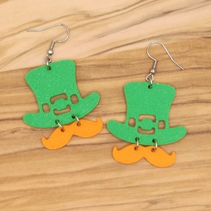 May include: A pair of green glitter leprechaun hat earrings with orange glitter mustaches hanging from the bottom.