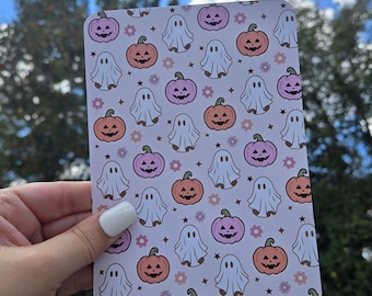 May include: A rectangular card featuring a repeating pattern of smiling jack-o'-lanterns in orange and pink, white ghosts, and small flowers. The background is a light lavender color. The card is held in a hand against a backdrop of a blue sky and trees.