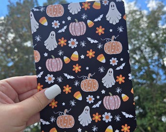 May include: A black rectangular card featuring a Halloween-themed pattern. The design includes orange pumpkins, pink pumpkins, white ghosts, candy corn, orange flowers, white flowers, bats, and stars. The card is held against a backdrop of a blue sky and green foliage.
