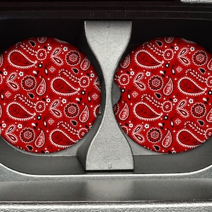 May include: Two car coasters with a red paisley pattern, featuring black and white accents. The coasters are circular and designed to fit in a car's cup holders. The paisley design includes teardrop shapes and floral elements.