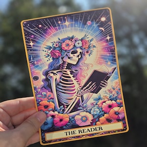 May include: A tarot card illustration featuring a skeleton figure wearing a floral crown and reading a book. The card has a colorful cosmic background with the words "THE READER" at the bottom. The card is held by a hand.