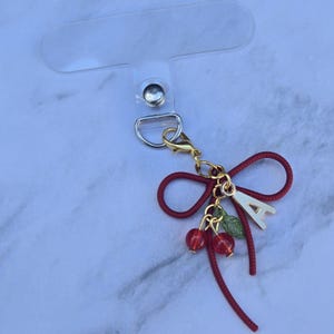 May include: A red bow-shaped phone charm with a gold-colored clasp and a clear plastic attachment. The charm features a gold-colored letter "A", red cherries, and green leaves. The charm is designed to attach to a phone.