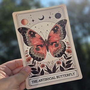May include: A rectangular card featuring a detailed illustration of a butterfly with red, black, and gray wings. The card has a celestial design with stars, a crescent moon, and the text "THE ANTISOCIAL BUTTERFLY".