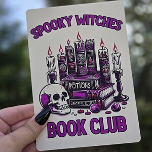 May include: A white card with purple text that reads "Spooky Witches Book Club". The card features an illustration of a skull, candles, and a stack of books with the words "Potions" and "Spells" on the spines.