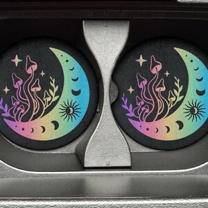 May include: Two round, black coasters with a rainbow crescent moon and mushroom design. The moon has a sun and crescent moon phases on it. The mushrooms are stylized and have a rainbow gradient.