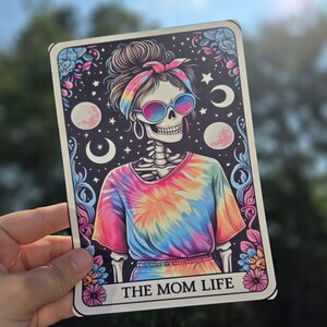 May include: A sticker featuring a skeleton wearing a tie-dye shirt and sunglasses. The skeleton has a bandana on its head and is surrounded by stars and moons. The text "THE MOM LIFE" is at the bottom of the sticker.