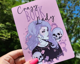 Puede incluir: Fondo rosa con una mujer de pelo morado que lleva una camisa negra y un collar con una cruz. La mujer sostiene dos cráneos. El texto "Crazy Book Lady" está escrito en morado en la parte superior de la imagen.