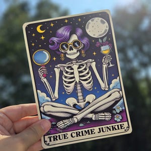 May include: A tarot card with a skeleton wearing purple hair, sunglasses, and earrings. The skeleton is holding a magnifying glass in one hand and a glass of wine in the other. The card has a dark blue background with stars and a crescent moon. The text "TRUE CRIME JUNKIE" is printed at the bottom of the card.