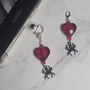 May include: Two red heart-shaped charms with black spider pendants. The charms are attached to silver-colored clasps. One charm is attached to a clear phone case. The word "kindle" is visible on the phone.