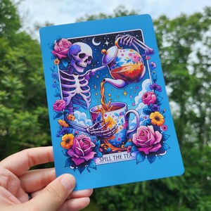 May include: A blue art print featuring a skeleton pouring tea into a floral teacup. The skeleton is surrounded by roses and flowers, with the text "SPILL THE TEA." The artwork has a whimsical, gothic theme.