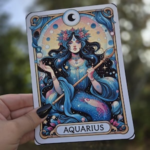 May include: A rectangular card featuring an illustration of a woman with long, dark hair, a floral crown, and a blue dress. The card has a gold border and the word "AQUARIUS" at the bottom. The woman is holding a staff with a mirror-like top.