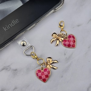 May include: Gold-toned heart-shaped charms with a pink and red checkered pattern and a gold bow. The charms are attached to a clear phone strap with a silver clasp. The word "kindle" is visible on the phone.