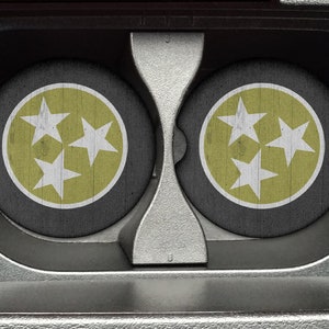 May include: Two round coasters with a black background and a white circle in the center. The circle contains three white stars on a yellow background. The coasters are in a cassette tape holder.