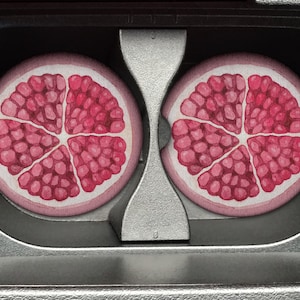 May include: Two pink and white coasters with a red pomegranate design. The coasters are in a cassette tape case.