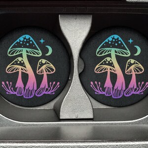 May include: Two round black coasters with a design of three rainbow mushrooms with white dots on top. The mushrooms are surrounded by small white flowers and a crescent moon with a star above it.