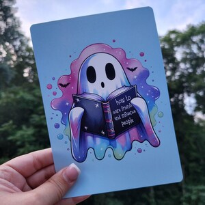 May include: A blue and purple ghost with a book that says "How to scare friends and influence people". The ghost is reading the book and has a surprised expression on its face. The background is a light blue color with white stars and bats.