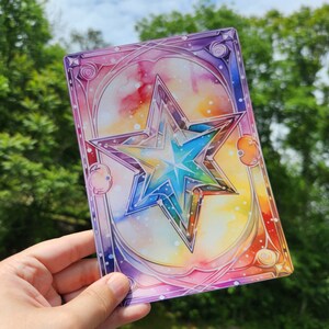 May include: A watercolor painting of a large, multi-colored star with a white center. The star is surrounded by a white, yellow, orange, pink, and purple background. The background has a white, speckled pattern. The star is framed by a decorative, ornate border.