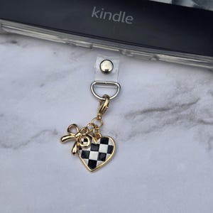 May include: A gold-colored charm with a bow and a checkered heart design. The charm is attached to a clear strap with a silver clasp, and the word "kindle" is visible in the background.