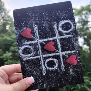 May include: A black chalkboard with a white tic-tac-toe game drawn on it. Three red hearts are placed on the board, two of which are in a winning row.