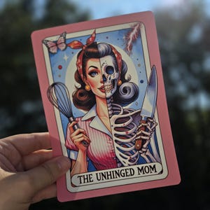 May include: A pink card with a cartoon illustration of a woman with a skeleton body. The woman is holding a whisk in one hand and a knife in the other. The card has the text "The Unhinged Mom" at the bottom.
