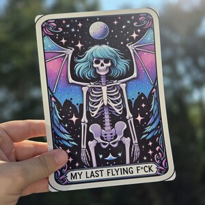 May include: A tarot card with a skeleton with wings against a dark blue background with stars. The skeleton has blue hair and is wearing a purple and pink outfit. The text on the card reads "My Last Flying F*ck".