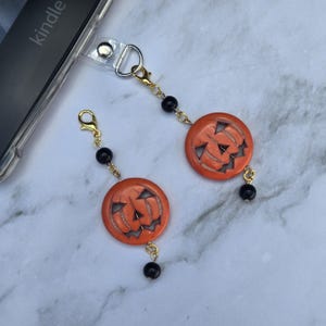 May include: Two orange pumpkin charms with carved faces, attached to gold-colored chains and black beads. The charms are connected to a phone case with a clear strap. The charms are a festive accessory for Halloween.
