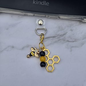 May include: A gold-colored bee charm with a yellow and black enamel bee, black and yellow beads, and a honeycomb charm. The charm is attached to a clear strap with a silver-colored metal clasp.