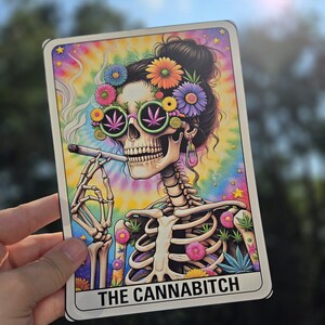 May include: A colorful illustration of a skeleton with a skull wearing sunglasses and a flower crown. The skeleton is holding a joint and is surrounded by flowers. The text "THE CANNABITCH" is at the bottom of the image.