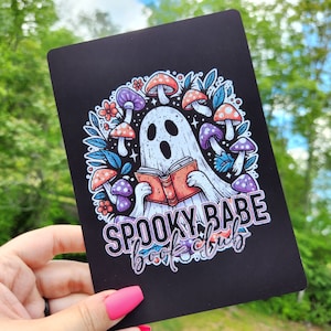 May include: A black card with a spooky illustration of a ghost reading a book surrounded by mushrooms. The text "Spooky Babe Book Club" is written in white with a pink outline.