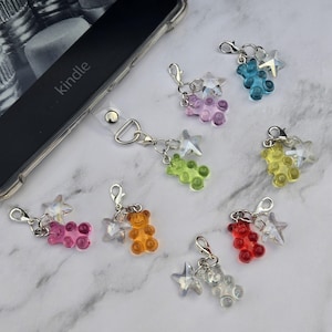 May include: A collection of colorful gummy bear charms with star accents and silver-tone clasps. The charms are in various colors, including pink, orange, green, blue, yellow, red, and clear. A clear phone case with the word "kindle" is visible in the background.