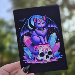May include: A black rectangular print featuring a purple cat with bat wings, sitting atop a skull with dripping pink liquid. The cat has a pentagram on its forehead and collar. Crystals and a crescent moon are in the background.
