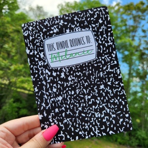 May include: A black and white composition notebook with the text "This  Windile Belongs To: Melanie" on the front cover.