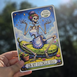 May include: A colorful illustration of a skeleton wearing a white apron and a floral crown sitting on a large green pickle. The text "Size Matters" is in a speech bubble above the skeleton's head. The text "In My Pickle Era" is at the bottom of the image.