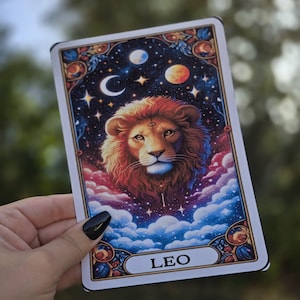 May include: A tarot card featuring a detailed illustration of a lion's head with a fiery mane, set against a cosmic background of stars, planets, and clouds. The word "LEO" is displayed at the bottom of the card.