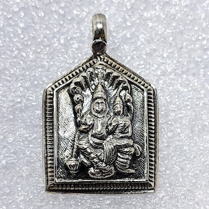 May include: Silver pendant featuring a detailed depiction of two deities, likely Hindu, with intricate carvings and a decorative border. The pendant is shaped like a house with a pointed roof.