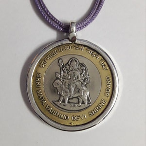 Op de afbeelding: Een zilveren en gouden ketting met een hanger met een afbeelding van een hindoeïstische godheid op een leeuw. De hanger heeft de tekst "Shri Mata Vaishno Devi Shrine Board" rond de rand.