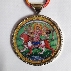 May include: A round pendant necklace with a gold-colored frame and a colorful image of a Hindu goddess riding a tiger. The image is surrounded by a decorative border.