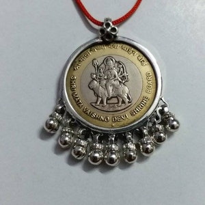 Puede incluir: Un colgante de plata con una moneda de oro que representa a una diosa hindú montando un león. La moneda tiene texto en hindi e inglés que dice "Shri Mata Vaishno Devi Shrine Board". El colgante está unido a una cuerda roja.