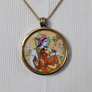 Puede incluir: Collar con colgante dorado. El colgante circular presenta una colorida ilustración de dos figuras con vestimenta tradicional. El colgante cuelga de una delicada cadena con un cierre. La ilustración representa a un hombre y una mujer en un abrazo amoroso.