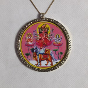 Puede incluir: Collar con colgante dorado. El colgante redondo presenta una imagen religiosa con un borde decorativo y un fondo rosa. La imagen representa una deidad de múltiples brazos montada sobre un león. La cadena del collar es dorada.