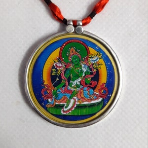 Puede incluir: Un collar con un colgante de plata redondo, con una imagen colorida de una deidad verde sentada sobre una flor de loto. El colgante está rodeado por un marco de plata y está unido a un cordón rojo y negro.