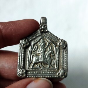 May include: Silver pendant with an intricate design featuring a figure riding a horse. The pendant is in the shape of a house with a pointed roof and has a loop at the top for hanging.