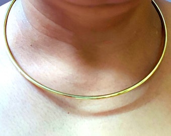 May include: A gold-colored choker necklace. The necklace is a simple, thin, and rigid band that sits close to the neck. The metal has a shiny, reflective surface.