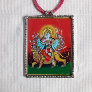 May include: A silver pendant with a colorful image of a Hindu goddess with multiple arms riding a lion. The pendant is on a red cord.