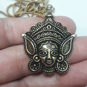 May include: A gold-coloured pendant necklace with a detailed face design. The pendant has an ornate, decorative crown and is suspended from a delicate chain. The face has detailed features against a white background.