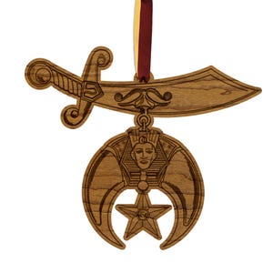 May include: Wooden decorative ornament featuring a saber, a crescent moon with a star, and a pharaoh's head. The ornament is suspended by a maroon and gold ribbon. The design is etched into the wood.