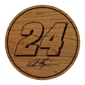 May include: A round wooden coaster with a dark brown border. The number "24" is prominently displayed in a bold, stylized font. Below the number is a signature. The wood grain is visible.