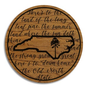 May include: A round wooden sign with a dark wood grain finish. The sign features a black outline of the state of North Carolina, a tree, and the text "Here's to the land of the long leaf pine...the Old North State."
