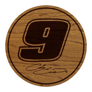 May include: A round wooden coaster with a dark brown number 9 in the center. The number is outlined in a lighter brown. Below the number is a signature in a cursive font. The coaster has a light brown wood grain.