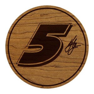 May include: A round wooden plaque with a dark brown number 5 in the center. The number has a shadow effect. A signature is in the lower right corner. The plaque has a light brown wood grain.